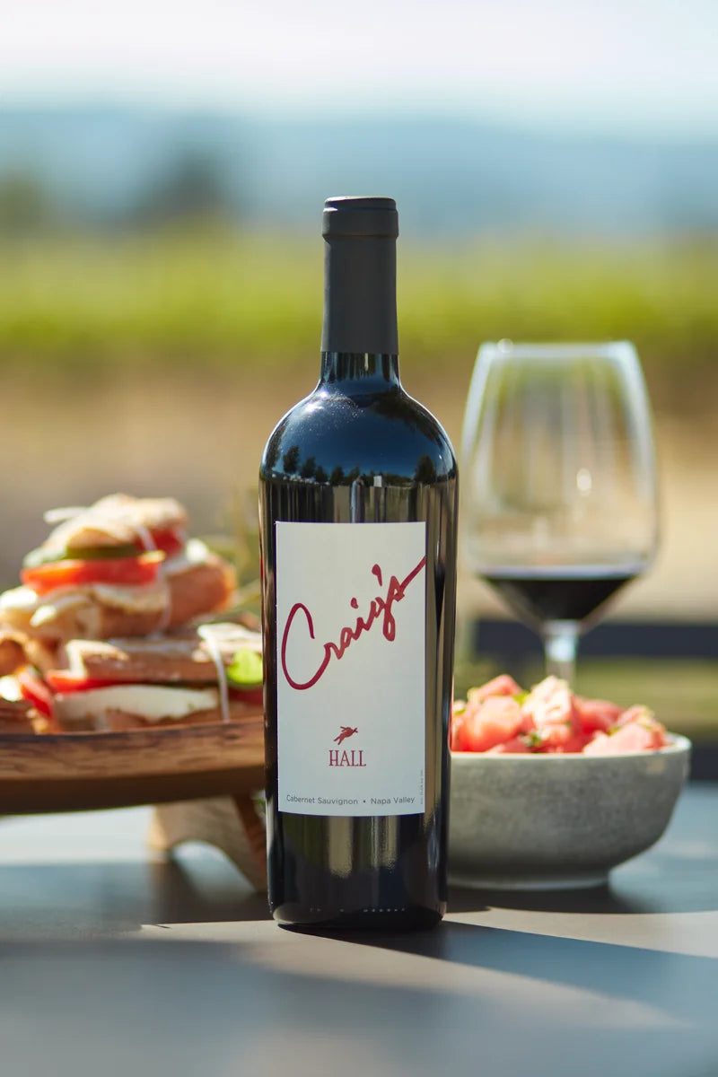 Photo of a bottle of 2023 HALL Craig's Cabernet Sauvignon on a picnic table with a platter of sandwiches, a summer salad, and glass of red wine in the background.