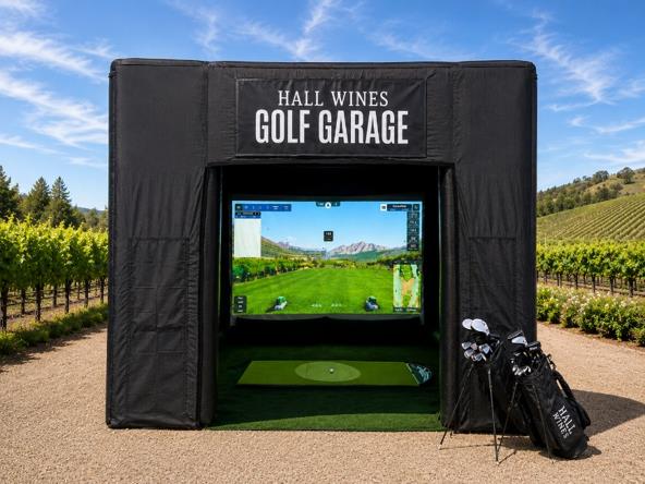 A black Hall Wines Golf Garage simulator tent set up outdoors on a gravel path, with a large screen showing a golf course, a green hitting mat inside, and golf clubs in a bag beside it. Vineyard rows and hills are visible in the background under a blue sky.
