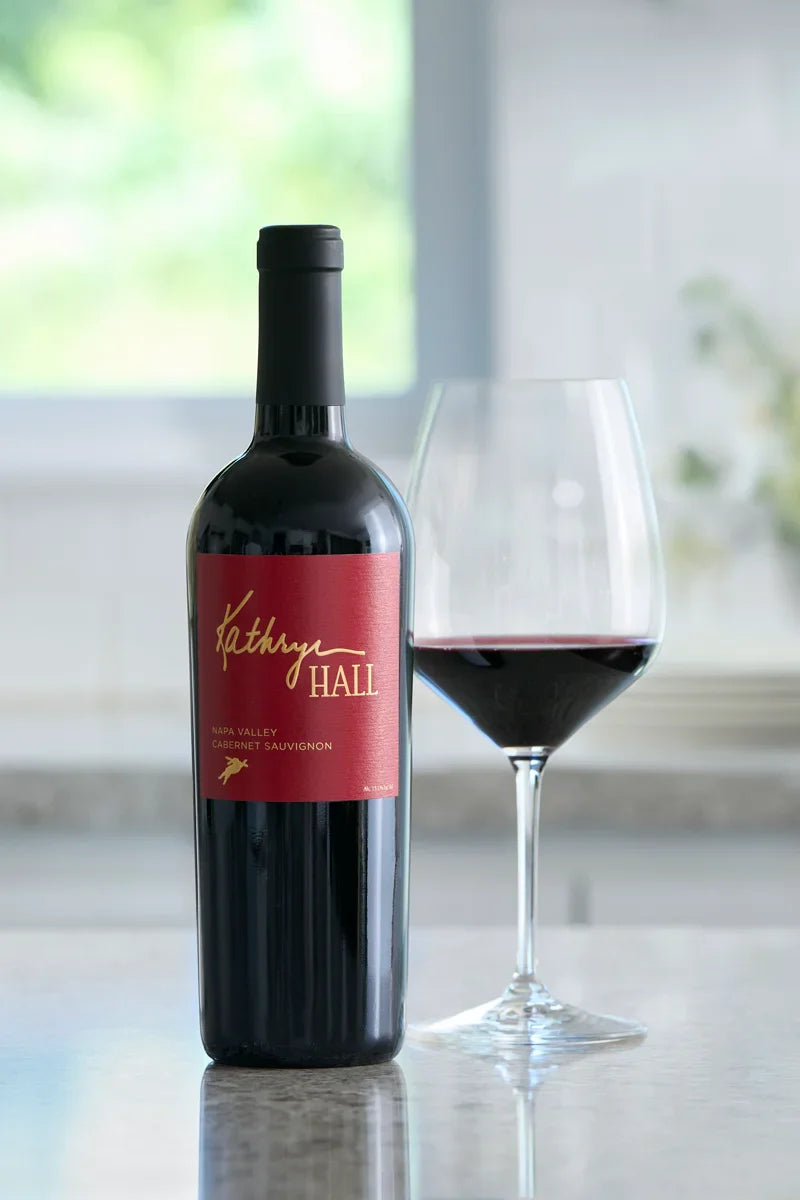 A bottle of Kathryn Hall Cabernet Sauvignon on a granite countertop next to a glass of red wine.