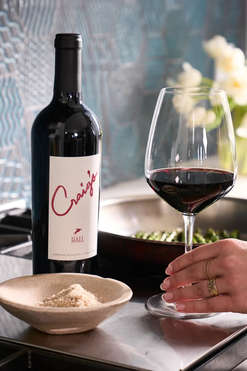 Woman reaching for a glass of red wine next to a bottle of 2020 HALL Craig's Cabernet Sauvignon, with a dish of shredded Parmesan and sautéing vegetables on the stove nearby