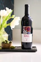 Image of a bottle of 2022 HALL Coeur Cabernet Sauvignon next to a small dish of olives with a vase of flowers in the background.