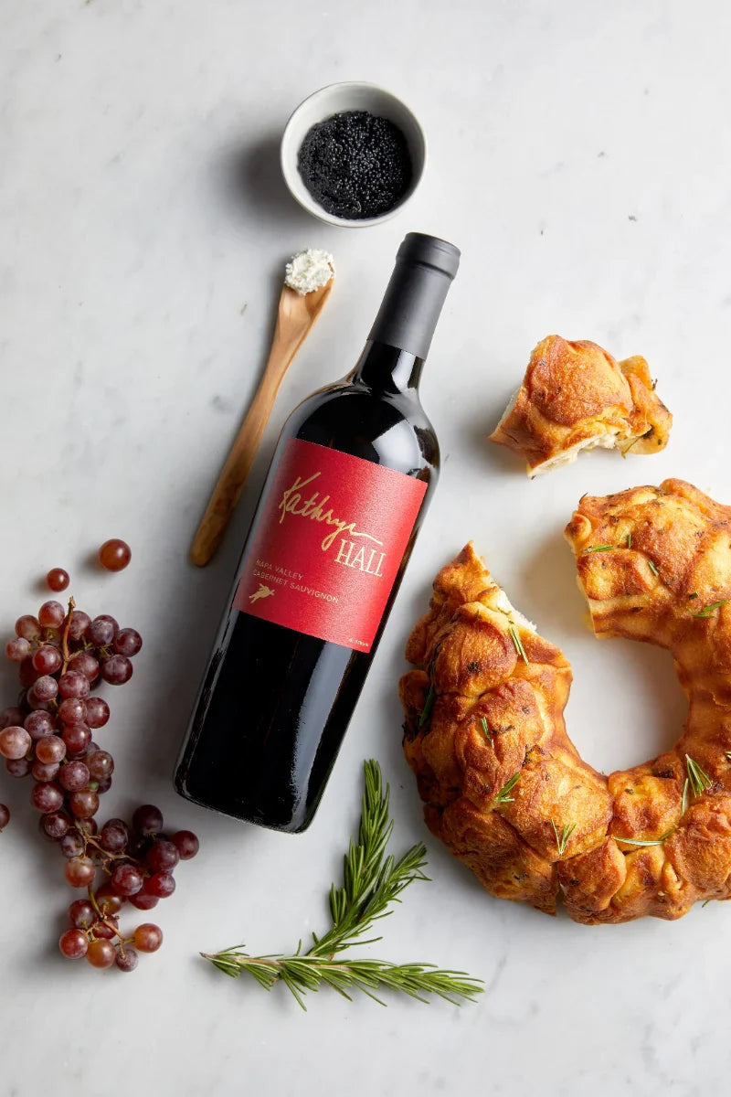 Image of a Kathryn Hall Napa Valley Cabernet laying flat on a marble counter surrounded by herbed bread, caviar, red grapes and creme fraiche.