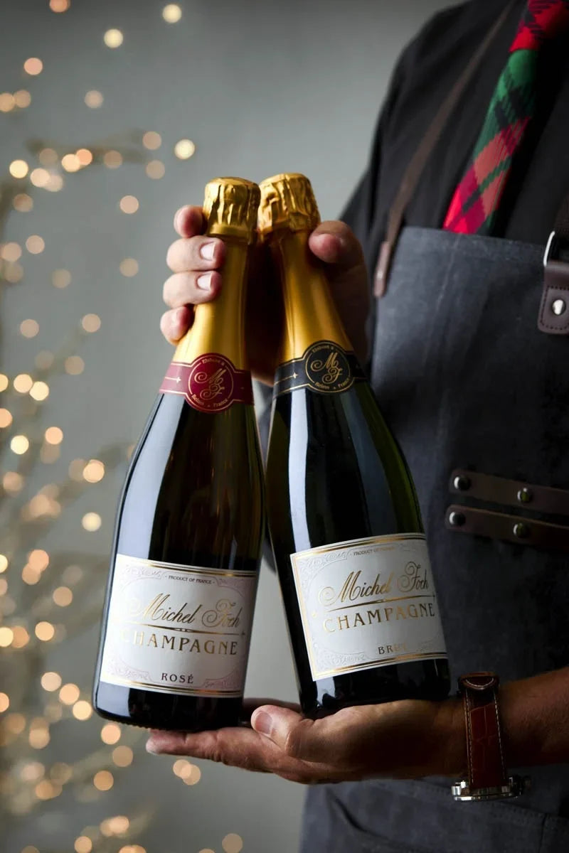 Image of the Michel Foch Brut & Rosé Duo gift set. The image is of a man wearing an apron and holiday tie holding the two bottles of Champagne included in this gift set with twinkling lights in the background.